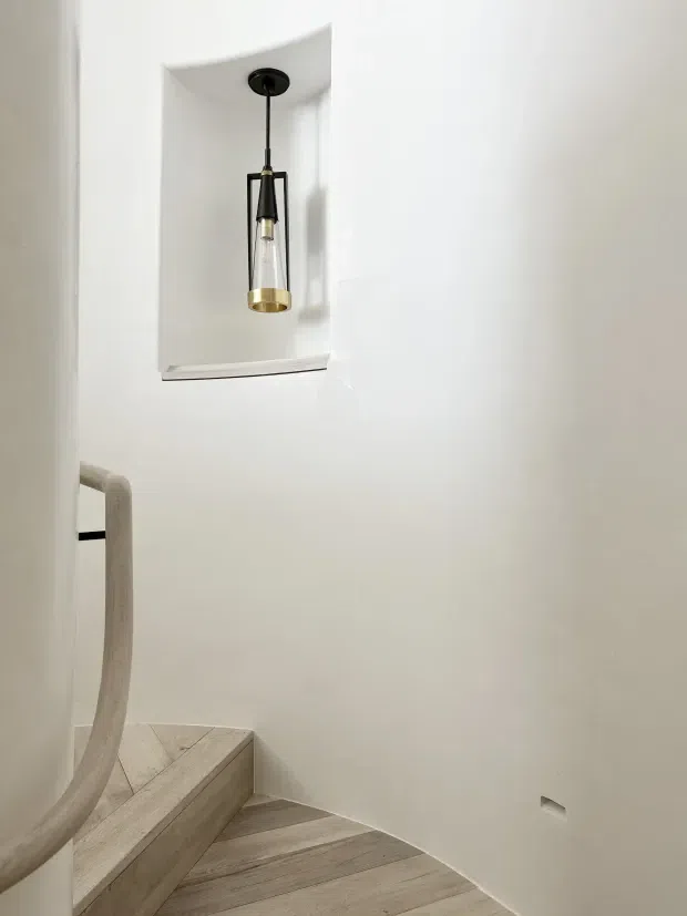 Minimalist interior stairway with pale timber herringbone flooring and matching stair treads, smooth white walls, a curved plaster recess, and a single suspended pendant light with black and brass detailing. A sculptural pale handrail follows the stair curve, creating a clean, refined architectural composition.