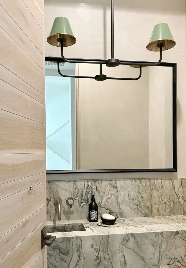 A refined bathroom vanity viewed through a partially open timber door. The space features a large black-framed rectangular mirror, a sculptural two-light pendant with pale green shades and dark metal arms, and a wall-mounted stone vanity with an integrated basin and matching heavily veined grey-and-white stone backsplash. The overall design is contemporary and luxurious with soft neutral tones and strong natural stone patterning.