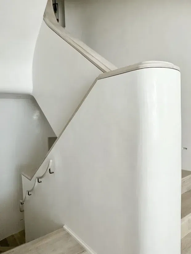 A close-up interior view of a curved stairway with a sculptural white plaster or painted balustrade, pale timber handrail, and light timber stair treads. The stair enclosure has smooth rounded corners and minimal detailing, with small dark hooks or fittings visible along the side.