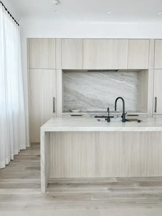 Bright, refined contemporary kitchen featuring a large island with a pale stone benchtop and vertically fluted light cabinetry. The kitchen includes integrated wall storage, a dramatic grey-and-white natural stone splashback, dark tapware, timber-look flooring, and full-height sheer curtains along the window wall.