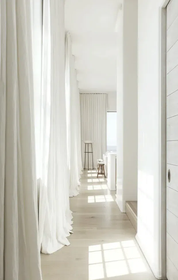 A bright, narrow white hallway or passage with full-height sheer curtains along the left side, pale timber flooring, white walls and trim, and strong rectangular sunlight patterns across the floor. A white horizontally panelled door with dark circular hardware is visible on the right, while the corridor leads toward a small stool and a bright window at the far end. The space has a clean, airy, minimalist appearance.