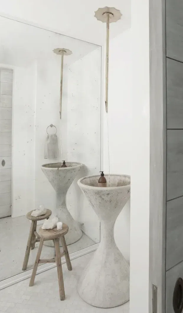 A minimalist bathroom featuring a sculptural freestanding vessel basin in a pale textured finish, suspended pendant lighting, a full-height wall mirror, and a small rustic wooden stool. The room is predominantly white with a light mosaic-tile floor and subtle grey detailing.