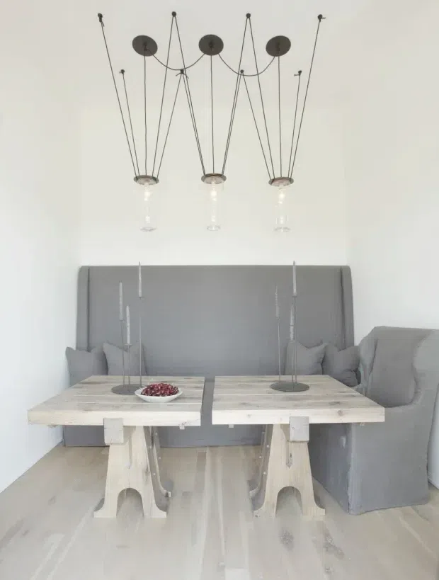 Bright, minimalist dining area with a pale timber floor and two rustic light-wood dining tables arranged together, paired with a long grey upholstered banquette and matching slipcovered dining chair. Three sculptural pendant lights with thin black suspension rods hang above the tables against white walls, while simple tall candleholders and a bowl provide restrained styling.