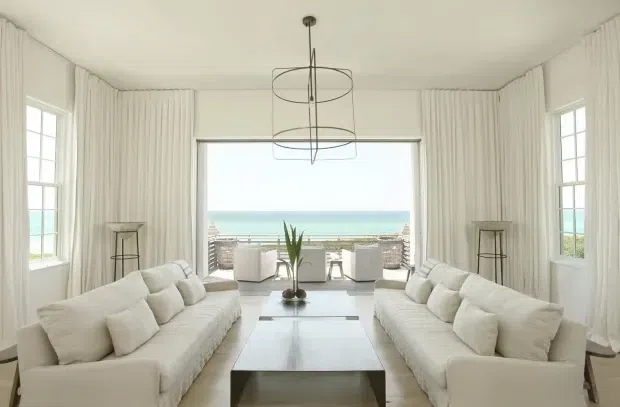 Bright, symmetrical coastal living room with two large white slipcovered sofas facing a low rectangular coffee table. Full-height white drapery frames broad ocean-facing glazing, with additional multi-pane windows on both sides. A minimalist circular pendant light hangs centrally, while pale flooring, neutral furnishings, and sparse decor create an airy, refined interior with a strong seaside outlook.