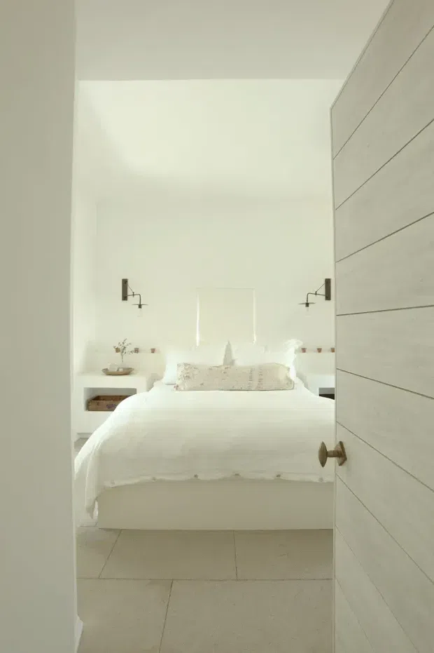 A bright, minimalist bedroom viewed through a partially open horizontal-panel door. The room features a white upholstered bed with layered white bedding and a textured neutral lumbar cushion, simple white bedside tables, small decorative objects, and matching wall-mounted reading sconces. Large pale floor tiles and white walls create a clean, airy atmosphere, while the door adds a light timber-toned feature.