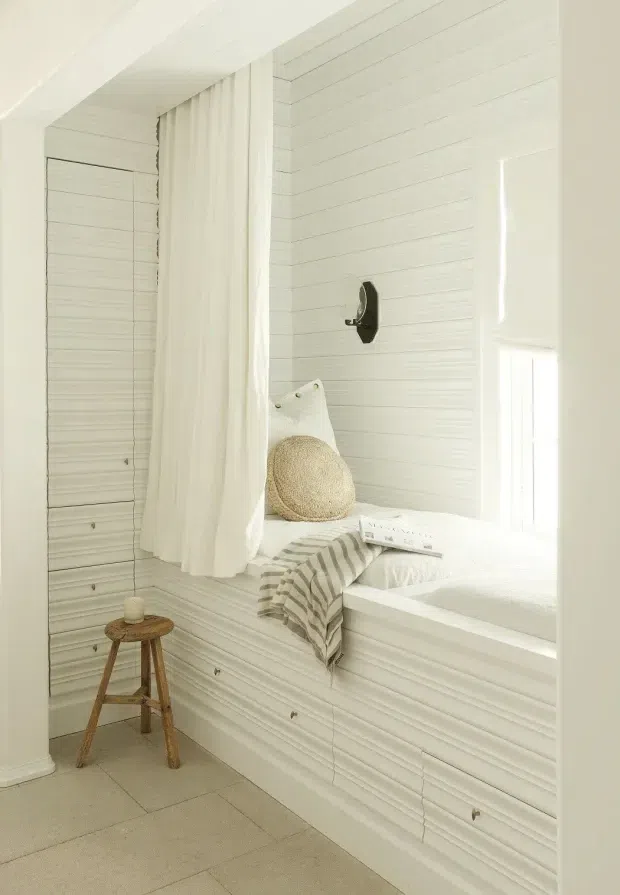 Bright, serene built-in window seat or daybed nook clad in white horizontal wall panelling. A sheer white curtain hangs beside the deep window recess, with layered white cushions, a round woven cushion, and a striped throw. A small rustic wooden stool sits on the pale tiled floor, while a dark wall-mounted reading light provides contrast.