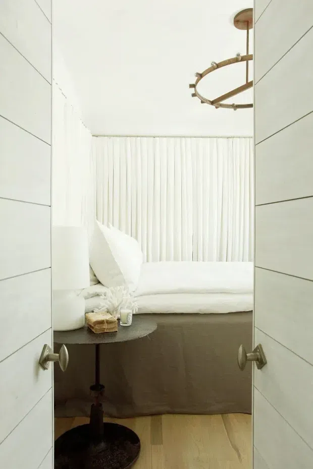 A softly lit bedroom viewed through partially open white paneled doors. The room features a low bed with white bedding, sheer floor-to-ceiling curtains, light timber flooring, a dark round side table, and a circular brass-toned pendant light. The overall design is minimal, monochromatic, and serene.