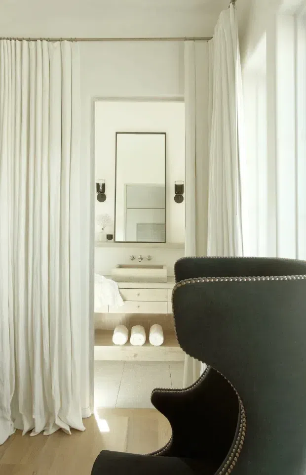 A bright, refined bedroom interior viewed through floor-length white curtains. A black wingback chair with nailhead trim dominates the foreground, while the background shows a neatly made bed, a pale built-in vanity or dressing table with drawers, a large rectangular mirror, wall sconces, and rolled towels. Pale timber flooring, white walls, and soft natural light create an elegant, serene atmosphere.