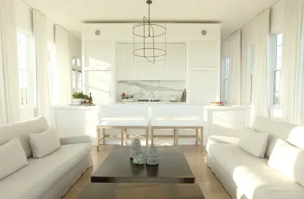 Bright, symmetrical white living room with two facing slipcovered sofas, a central dark rectangular coffee table, white built-in cabinetry, a marble-look feature panel and fireplace or display niche, and a dining area with bench seating beyond. Tall multi-pane windows are dressed with sheer curtains, and two minimalist circular pendant lights hang from the ceiling. The room has a refined coastal-traditional feel with abundant natural light and pale timber flooring.
