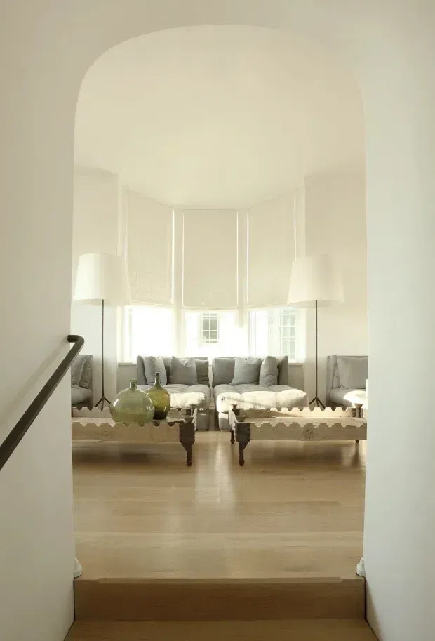 A bright, serene living room viewed through a wide rounded archway from a short stair or entry landing. The room features pale timber flooring, white walls and ceiling, a large bay of tall windows with white blinds, a light upholstered sofa and matching ottomans, slender floor lamps, and muted green glass vessels on a rustic low table. The overall composition is soft, airy, and neutral with understated traditional detailing.