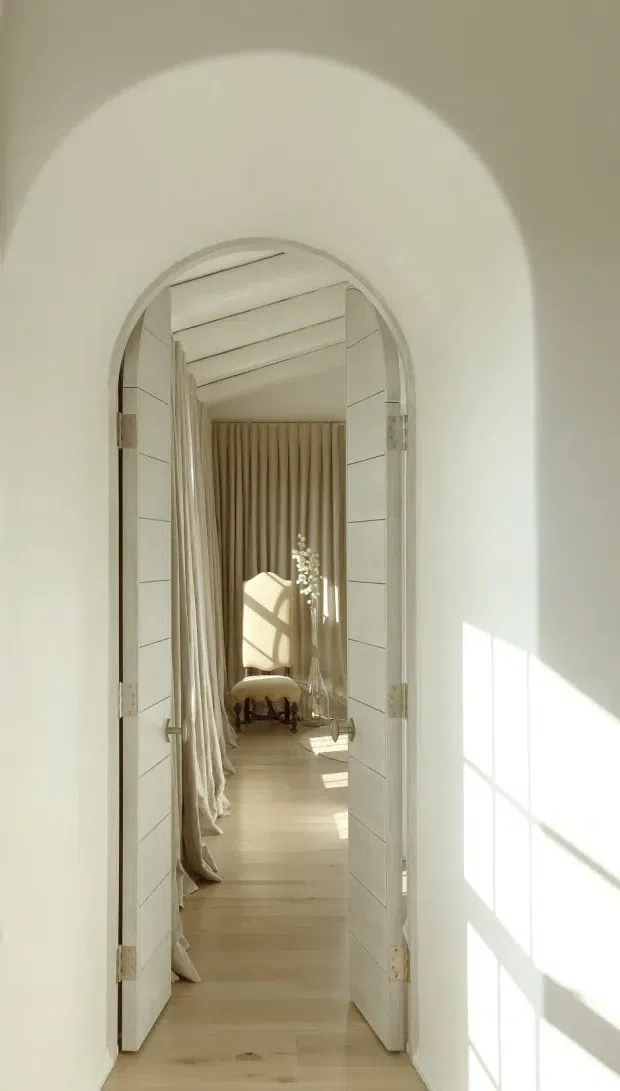 A softly lit interior hallway viewed through an arched, white paneled double doorway. The passage has pale timber flooring, white walls, full-height beige curtains, and a glimpse of a refined sitting area with an upholstered chair and small stool at the far end. Strong sunlight casts geometric window shadows across the walls and floor, creating a warm, serene atmosphere.