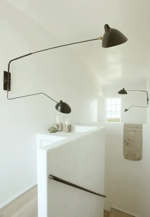 Bright minimalist interior corner featuring sculptural, articulated dark metal wall lamps mounted on white walls and a white built-in partition or stair balustrade. A small multi-pane window, pale timber flooring, simple decorative objects, and a muted wall hanging are visible. The space has a clean, restrained contemporary feel with strong white surfaces and high natural light.