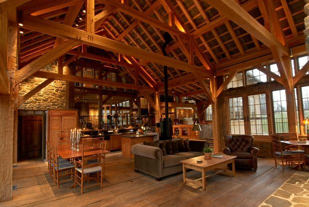 A spacious rustic lodge-style open-plan interior with exposed timber post-and-beam framing, a high pitched timber roof structure, stone walls, large multi-pane windows, and warm wood flooring. The room combines a kitchen and dining area with a comfortable living space furnished with sofas and leather armchairs around a timber coffee table. Warm ambient lighting and natural materials create a cozy, inviting atmosphere.