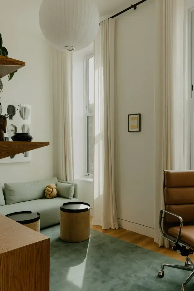 A refined, light-filled sitting and workspace corner with tall windows dressed in full-height cream curtains, pale walls, and warm timber flooring. A pale green sofa sits beside cylindrical coffee tables, while a tan leather office chair occupies the right side. Built-in or wall-mounted timber shelving displays ceramics and decorative objects, and a large white sculptural pendant hangs overhead. The room has a calm, contemporary mid-century-inspired feel with layered natural textures and muted colours.