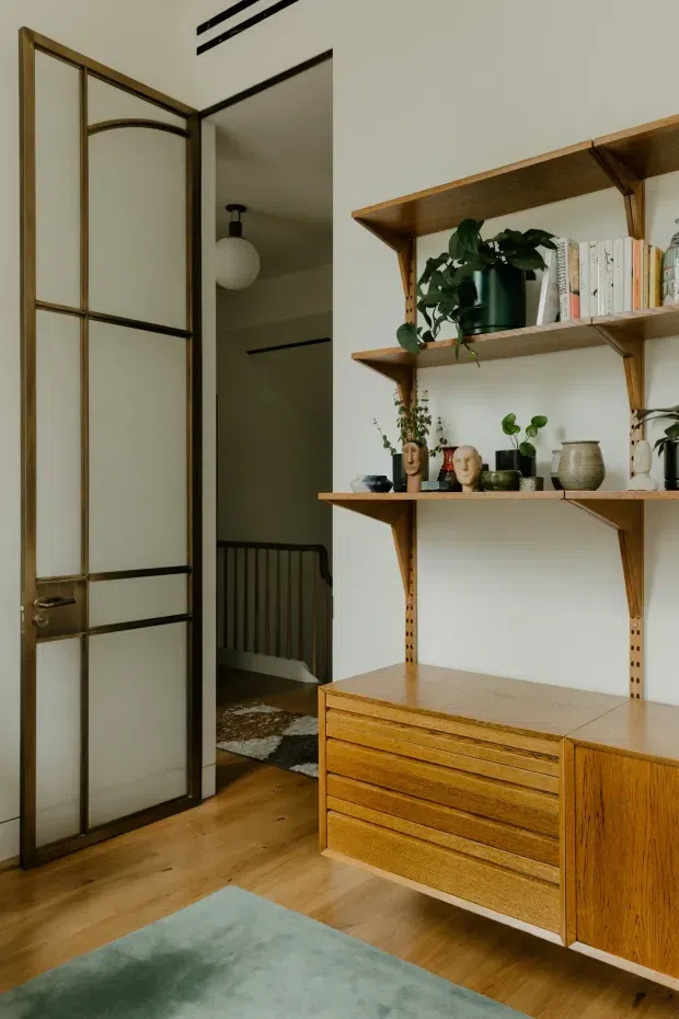 A warm, carefully styled interior corner featuring a tall glazed timber-framed door opening to a hallway and stair, with a wall-mounted timber shelving and drawer unit. The shelving displays books, potted plants, ceramics, and sculptural objects. Timber flooring, white walls, dark metal door detailing, and a muted blue-green rug create a refined mid-century-inspired atmosphere.