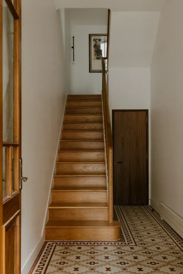 A narrow residential hallway and stairway with warm timber stairs, a matching dark timber door and handrail, cream painted walls, and patterned geometric mosaic tile flooring. A glazed timber entry door is visible at left, with framed artwork at the stair landing. The space has traditional detailing and a warm, refined character.