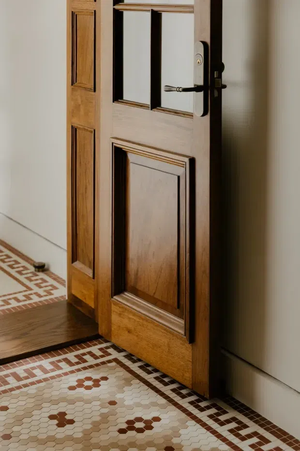Close-up interior view of a partially open, richly detailed timber door with raised decorative panels, dark trim, and a dark metal lever handle. The doorway opens onto a patterned geometric mosaic tile floor in cream, beige, brown, and terracotta tones, with a dark timber floor visible beyond. Plain light-coloured walls frame the traditional door joinery.