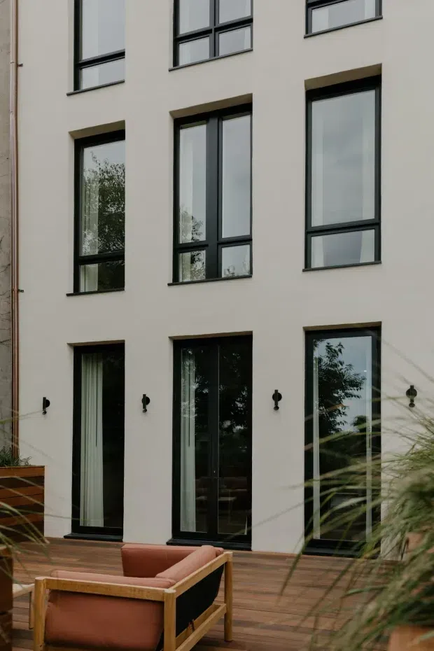 Exterior view of a contemporary multi-storey residential building with a pale rendered facade, regularly arranged black-framed windows and full-height glazed doors. A timber deck with outdoor lounge seating occupies the foreground, with planting partially screening the lower right. Reflections reveal surrounding trees in the glazing.