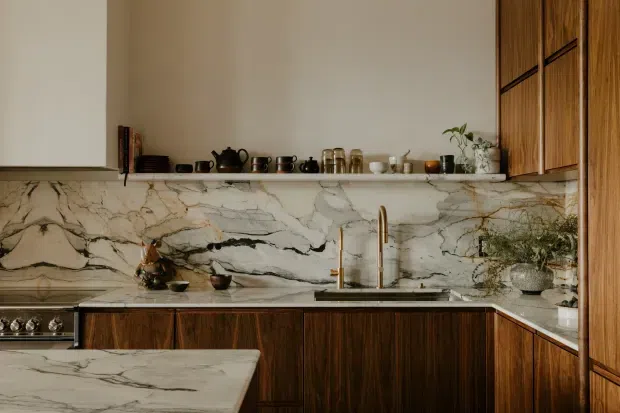 A refined contemporary kitchen featuring dark timber cabinetry, a dramatic veined marble backsplash and matching waterfall-edge island, brass-toned fixtures, open display shelving, and integrated appliances. Warm natural materials and curated pottery create a luxurious, calm atmosphere.