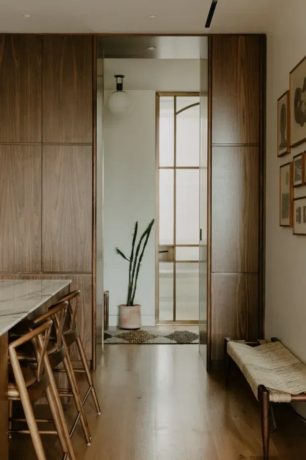 A warm, refined interior view of a compact transitional passage beside a kitchen or dining area. Full-height dark timber cabinetry lines both sides of the space, framing a glazed timber-framed doorway and a small entry or bathroom zone beyond. A stone island with timber bar stools is visible at left, while a woven bench sits along the right wall beneath framed artwork. The space has polished timber flooring, soft neutral walls, restrained lighting, and a tall potted plant.