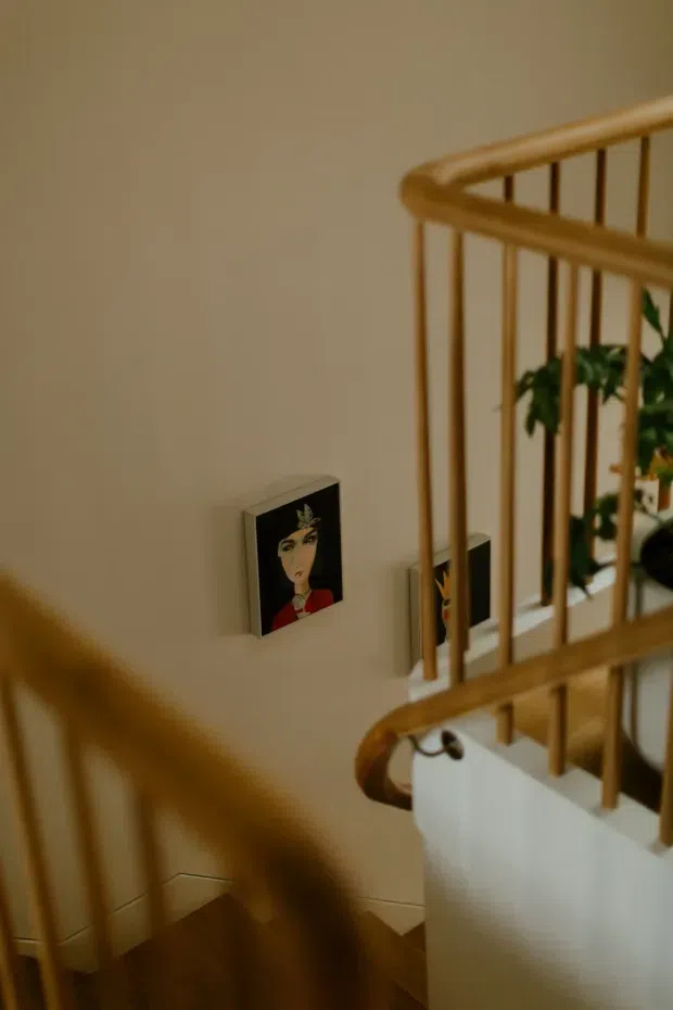 A warmly lit interior stairway viewed from above, featuring honey-toned timber handrails and vertical balusters, beige walls, and dark timber stair treads. Small framed artworks and a leafy plant are visible along the stair landing.