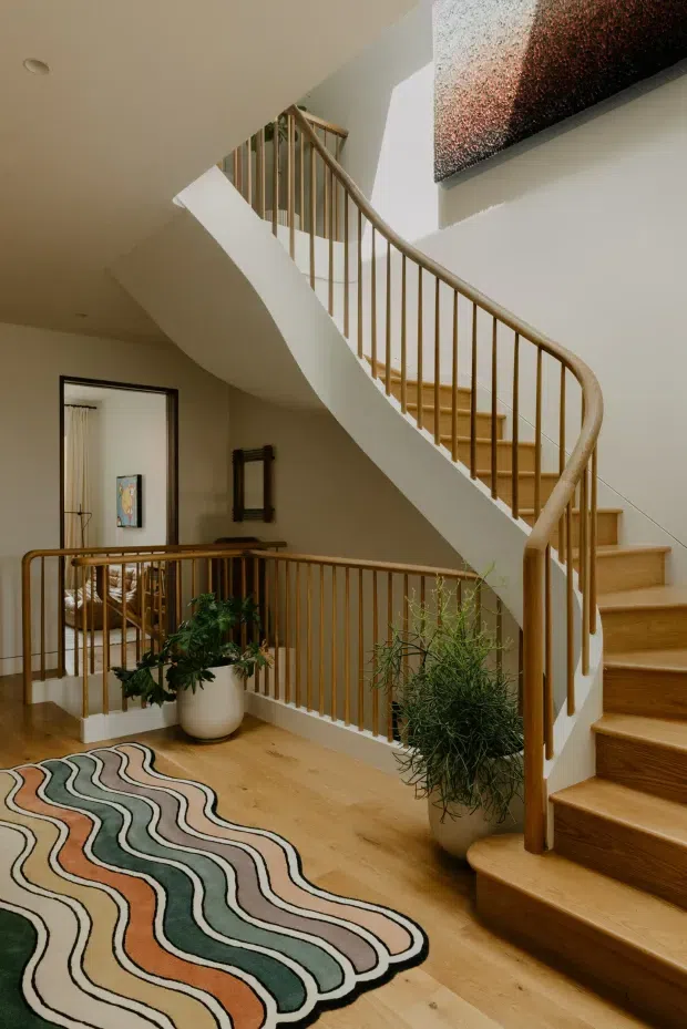 Interior view of a curved timber staircase and balustrade in a warm, multi-level hallway or landing. The staircase has white sculptural stringers, natural timber treads and handrails, and slender vertical balusters. A timber floor, abstract wavy-patterned rug, potted plants, framed artwork, and a glimpse of a bedroom or sitting area are visible. The space is softly lit with neutral walls and a large textured artwork overhead.