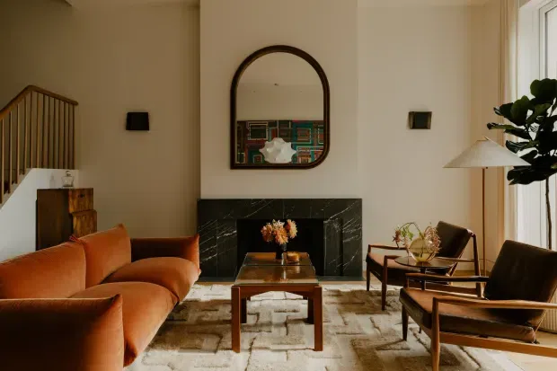 Warm, refined living room with a large rust-orange sectional sofa, two mid-century-style leather lounge chairs, a timber coffee table, and a black marble fireplace surround. An arched dark-framed mirror hangs above the fireplace, reflecting colourful artwork or shelving. The room has cream walls, a textured neutral rug, a sculptural floor lamp, indoor greenery, and abundant natural light from tall windows. A stairway is visible at left.