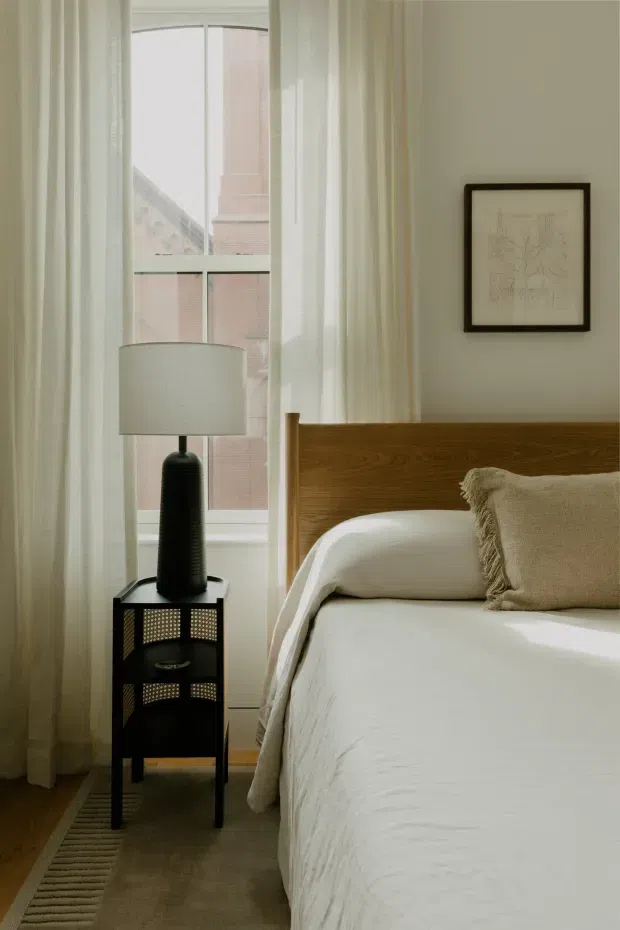 A softly lit bedroom corner with a timber bed featuring a warm medium-brown headboard, crisp white bedding, and a textured beige throw pillow. A black bedside table with cane-panel detailing supports a dark lamp with a pale fabric shade. Full-height sheer curtains frame a tall window, and a small framed artwork hangs on the light neutral wall. The room has a calm, refined, contemporary-traditional feel.