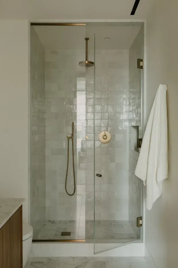 A compact walk-in shower enclosure in a refined bathroom, framed by clear glass doors and lined with glossy pale stone-look rectangular tiles. The shower features brushed brass/gold fixtures, including a ceiling-mounted rain shower, handheld shower, controls, and hinges. Marble-look flooring continues into the bathroom, with a timber vanity and partial toilet visible at the left edge. A white towel hangs on the right wall.