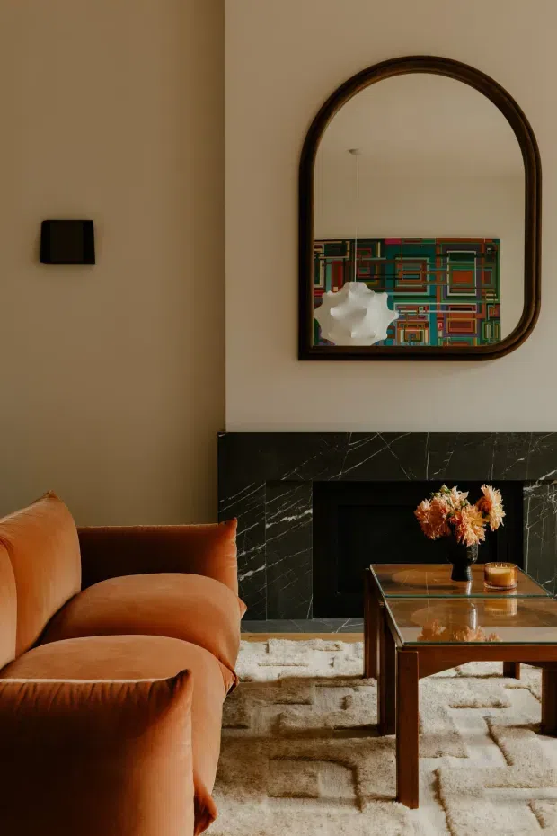 A warm, refined living room centered on a black marble fireplace with a large arched dark-framed mirror above it. A rust-orange upholstered sofa and matching seating occupy the foreground, paired with a glass-topped wooden coffee table and a textured cream rug. The mirror reflects a colourful geometric artwork and a sculptural pendant light, while dried flowers add a small decorative accent.