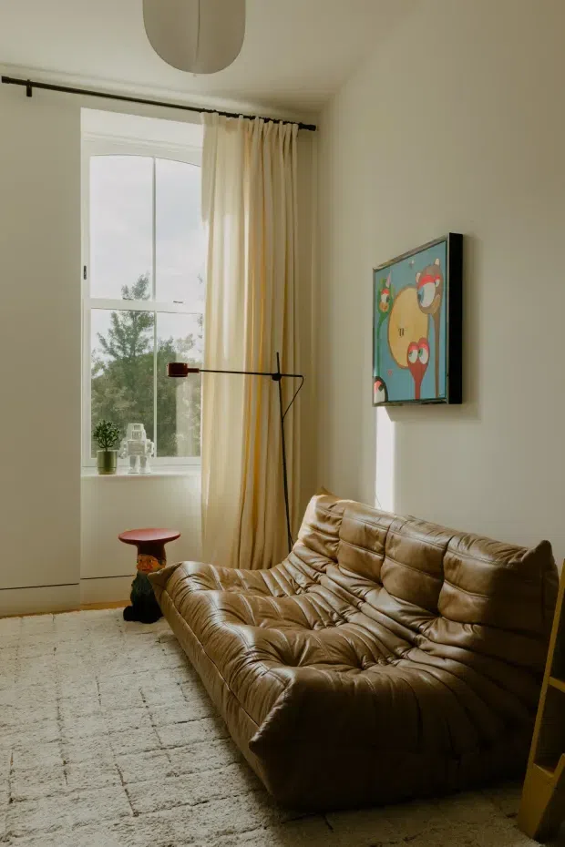 A warm, softly lit living room with a large brown tufted leather sofa, cream textured rug, tall window with sheer cream curtains, slim black floor lamp, framed colourful artwork, and a small side stool. The room has pale neutral walls, white trim, and a simple ceiling light, creating a vintage-inspired, cozy interior.