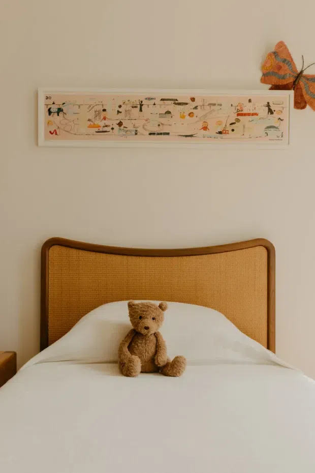 A warm, softly lit bedroom vignette centered on a neatly made bed with a curved timber-framed, woven cane headboard and white bedding. A teddy bear sits against the pillows. The pale neutral wall is decorated with a long framed illustrated artwork and a partial orange textile wall decoration, creating a gentle, playful and understated atmosphere.