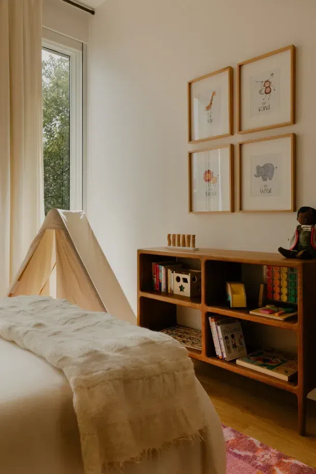 Warm, softly lit children's bedroom with a low bed covered in a cream textured throw, a small fabric play tent beside a tall window, and a mid-century-style wooden open shelving unit filled with books, toys, and decorative items. Four framed animal illustrations hang above the shelving, and a small doll sits on top. Light timber flooring, neutral walls, sheer curtains, and a pink patterned rug create a calm, playful atmosphere.