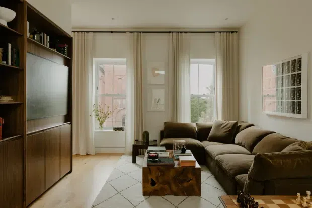 Warm, refined living room with a large dark timber built-in media wall and shelving, a deep neutral sectional sofa, pale timber flooring, full-height cream curtains, and a sculptural timber coffee table. Tall windows bring in soft natural light, while framed artwork and a chess set add understated character.