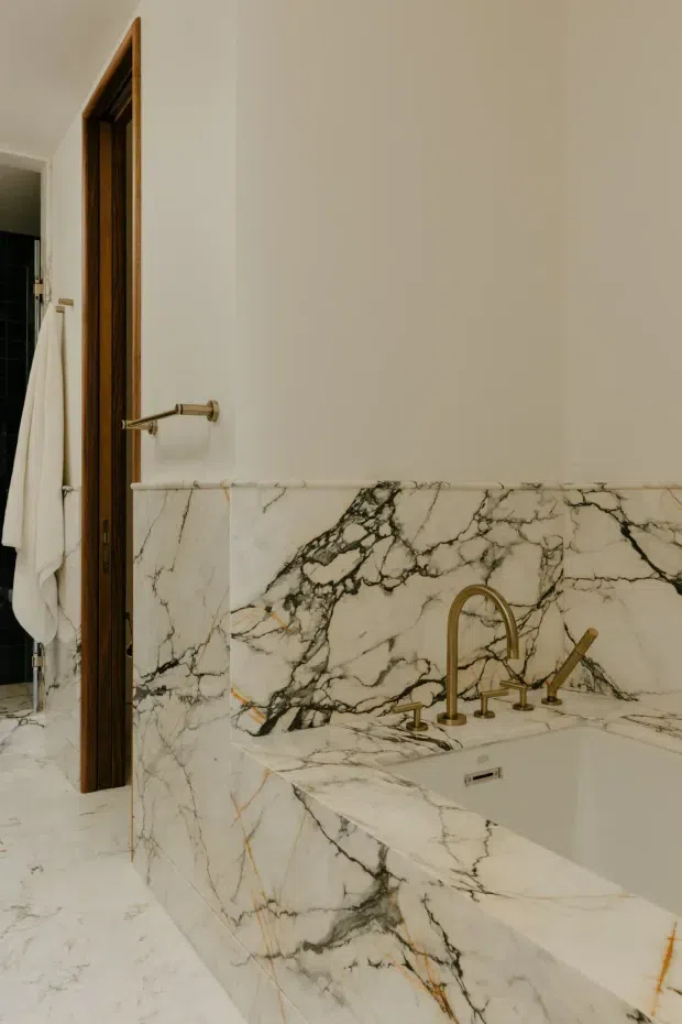 A refined bathroom featuring a built-in bathtub and surrounding walls clad in dramatic white marble with bold black and grey veining. A brass-toned wall-mounted bath filler and hand shower provide a warm metallic accent. The room has warm white painted walls, pale marble flooring, and a dark timber door frame, with a towel and part of an adjacent shower area visible at left.