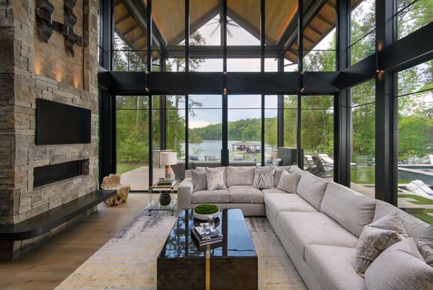 A spacious contemporary living room with a large L-shaped sectional sofa, black reflective coffee table, and stone-clad fireplace wall with a mounted television. Floor-to-ceiling black-framed glazing surrounds the room, opening views toward a wooded waterfront setting and outdoor seating. The dramatic pitched ceiling is lined with warm timber, while exposed dark structural framing and extensive glazing create a bright, refined interior.