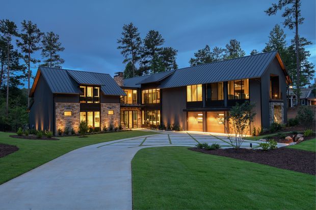 Twilight exterior view of a contemporary multi-wing residence set among tall trees. The home features dark vertical cladding, extensive warm-lit glazing, steeply pitched standing-seam metal gable roofs, and stone-clad sections. A broad curved concrete driveway and carefully landscaped lawns lead to the attached garage and glazed entry connection.