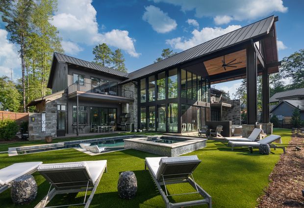 Contemporary multi-level residential home with dramatic pitched metal roofs, extensive floor-to-ceiling glazing, dark structural framing, and stone-clad walls. The house opens onto a landscaped backyard with an artificial-turf lawn, rectangular swimming pool, raised square water feature or spa, and multiple outdoor loungers. A covered outdoor area features a timber-lined ceiling and large ceiling fan. Mature trees and neighbouring homes surround the property.