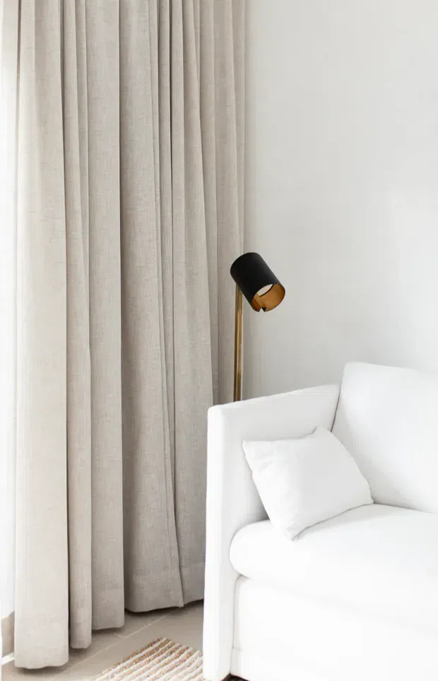 Bright, minimalist interior corner featuring a white upholstered sofa with a white cushion, full-height sheer beige curtains, a slim brass-and-black floor lamp, and a small neutral striped rug. The palette is predominantly white, cream, and light beige with soft natural light and very little visual clutter.
