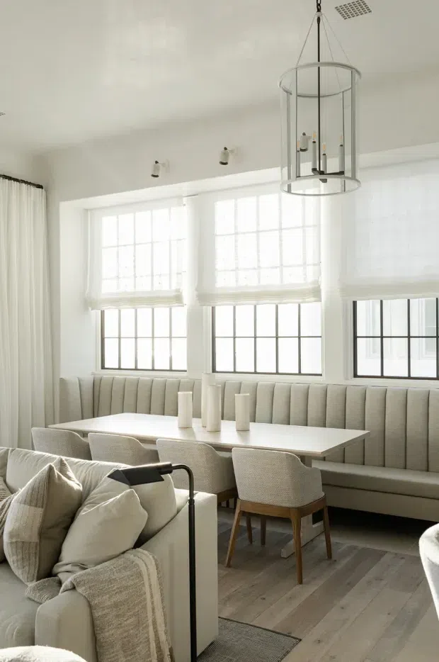 Bright, refined dining and living area with a built-in upholstered banquette beneath a bank of black-framed windows. A pale rectangular dining table is surrounded by upholstered chairs, while a light neutral sofa with cushions and a throw occupies the foreground. White walls, sheer curtains, layered Roman blinds, light timber flooring, and a large cylindrical pendant create an airy, understated interior.
