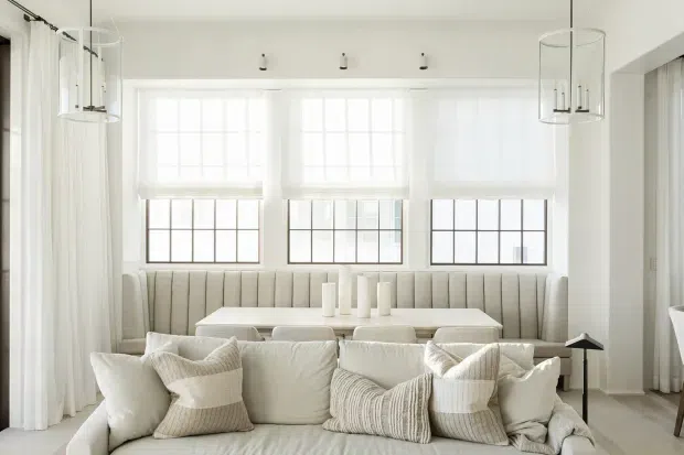 Bright, refined dining and living area with a built-in upholstered banquette beneath three large multi-pane windows. A white dining table sits behind a cream sectional sofa, with layered neutral cushions, sheer curtains, and cylindrical pendant lights creating an airy, softly textured interior.