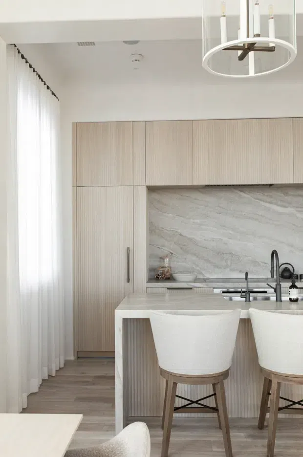 Bright, refined contemporary kitchen with a pale vertically fluted timber cabinetry wall, large light veined stone splashback, and a stone island with integrated sink and seating for two. Sheer full-height curtains, pale timber flooring, sculptural white pendant lighting, and upholstered timber bar stools create a soft neutral interior.