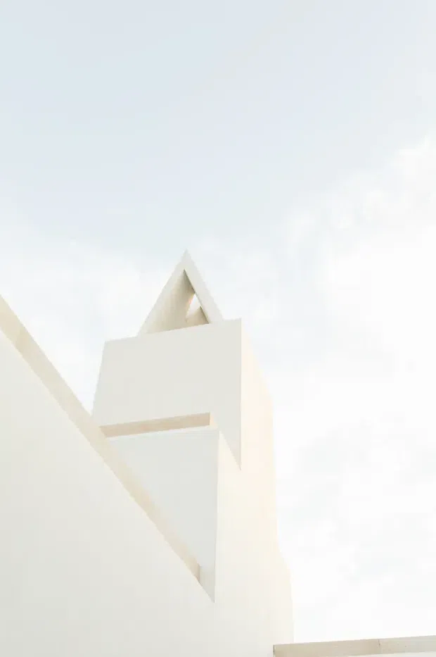 Minimalist architectural detail viewed from below, showing stark white geometric wall planes, a projecting horizontal element, and a triangular roof or clerestory opening against a bright overcast sky. The image emphasizes abstract form, sharp angles, and a highly restrained monochromatic facade.