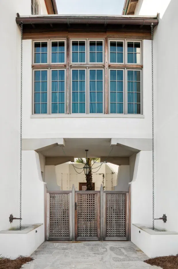 A narrow, enclosed courtyard passage framed by white rendered walls, with a two-storey timber-framed window arrangement above a covered entrance. The windows have pale timber frames and blue-green glazing or curtains, while weathered timber lattice gates close off the lower passage. Decorative rain chains descend from the roof gutter into built-in white basins on either side, and the floor is paved in irregular pale stone. The architecture has a distinctive courtyard, Mediterranean-influenced character with rustic timber detailing.