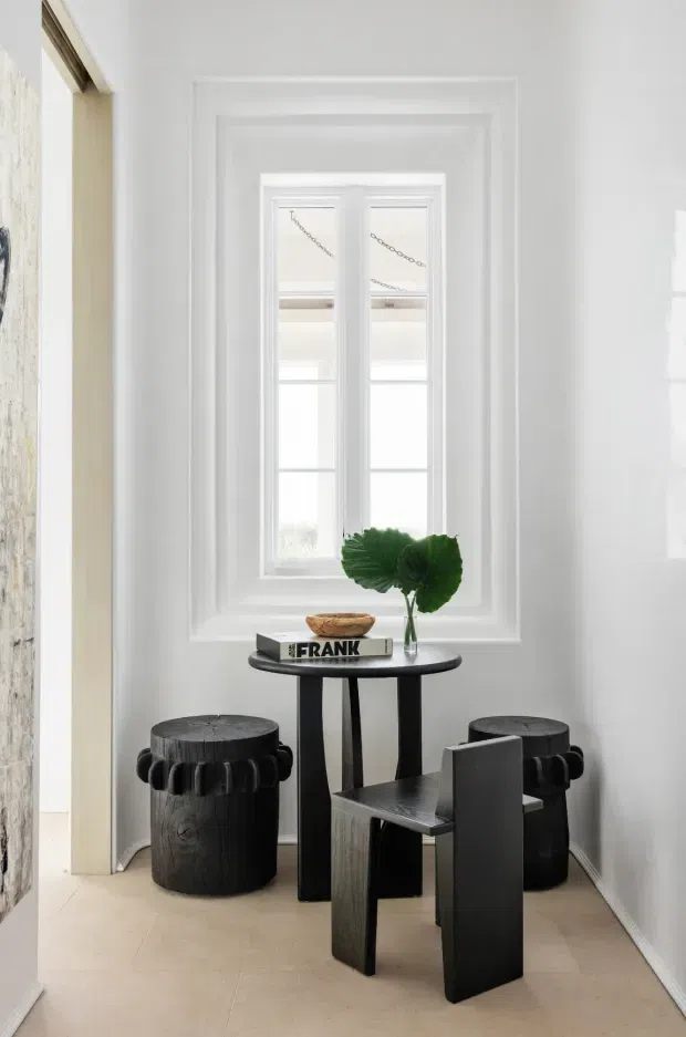 A bright, narrow interior nook styled as a small seating and display area. White walls and layered decorative window trim surround a tall, multi-pane window with white framing. A small black round table sits in front of the window with a vase of green leaves and books, accompanied by two dark cylindrical stools and a sculptural angular black chair. The pale floor and predominantly white walls create a clean, high-contrast setting with dark furniture accents.
