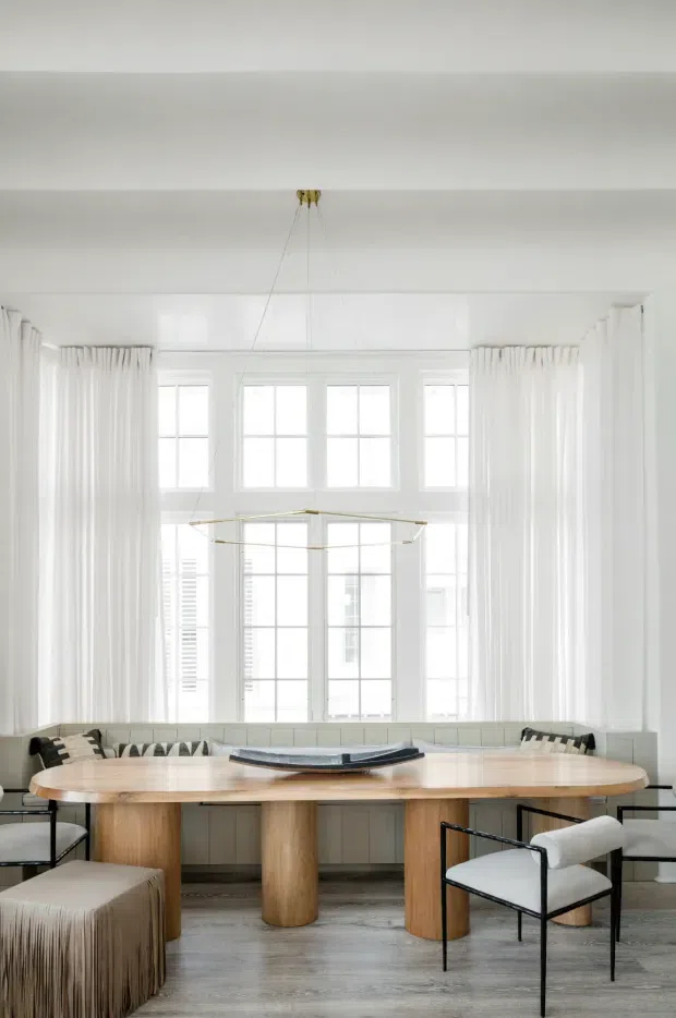 Bright, elegant dining room with a long rounded timber dining table, upholstered built-in banquette seating, black-framed dining chairs, and a sculptural linear pendant light. Full-height white curtains frame tall multi-pane windows, while pale timber flooring and white wall panelling create an airy, refined interior.