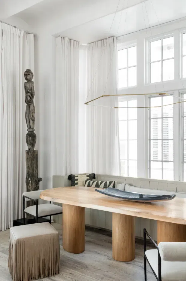 Bright, elegant dining area with a long light-wood dining table supported by cylindrical timber legs, upholstered dining chairs, a fringed ottoman, and a built-in upholstered banquette. White floor-to-ceiling curtains and large multi-pane windows create an airy setting, complemented by a sculptural pendant light and a tall carved wooden sculpture. The space has a refined, softly layered contemporary look with natural wood and neutral upholstery.