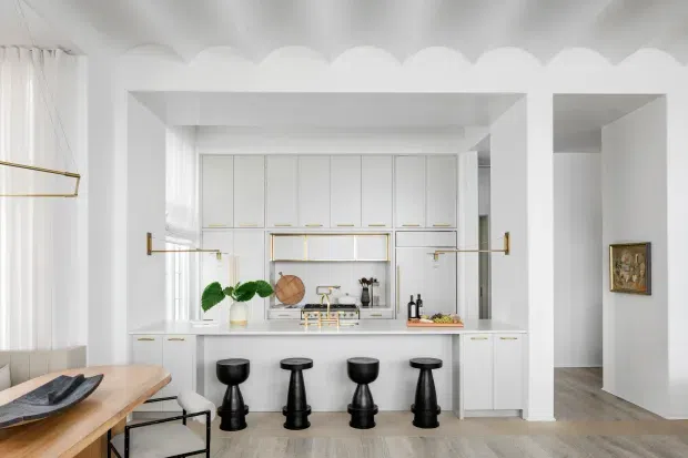 Bright white kitchen with a central island and four black bar stools, framed by a dramatic scalloped ceiling and deep square arch openings. The space features white paneled cabinetry with slim brass pulls and rails, a pale stone island benchtop, light timber flooring, sheer curtains, and restrained contemporary styling with brass accents.