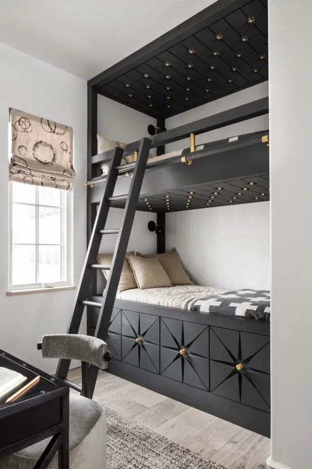 A compact, highly designed bunk-bed room with two stacked sleeping platforms in matte black, accessed by a leaning ladder. The built-in beds feature geometric, starburst-patterned drawer fronts with small gold knobs, while the undersides of the bunks are decorated with a grid of gold-accented star motifs. Neutral bedding, a pale timber-look floor, white walls, a patterned Roman blind, and a small upholstered desk chair complete the refined, dramatic space.