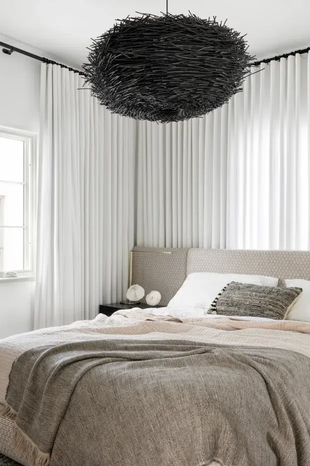 Bright, serene bedroom with a layered neutral palette. A large bed with a textured grey-beige throw, white bedding, woven cushions and an upholstered headboard sits against full-height white pleated curtains. A dramatic oversized black woven twig pendant light hangs centrally from the ceiling, with a small dark bedside table and sculptural bedside objects. The room has abundant natural light from a side window and black curtain hardware.